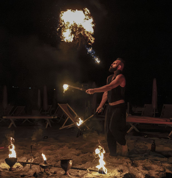 Uomo in spiaggia durante la notte mentre soffia una fiamma luminosa; performance di un mangiafuoco ripresa nel momento esatto dell’esplosione di fuoco. Man on a beach at night blowing a bright flame; firebreather performance captured at the exact moment of the fire burst.